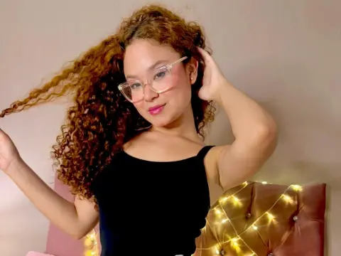 LunaBlush poses with her long curly hair and glasses, wearing a black tank top in a room with warm lights.