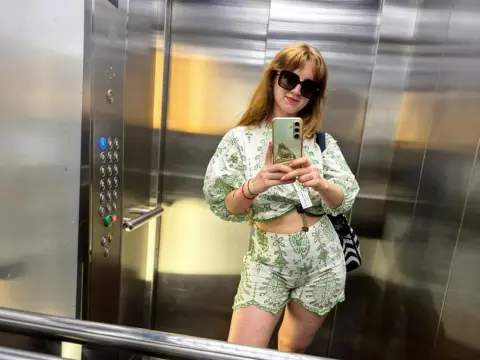 AnissaDolll takes a mirror selfie in an elevator wearing a green and white patterned crop top and matching shorts.