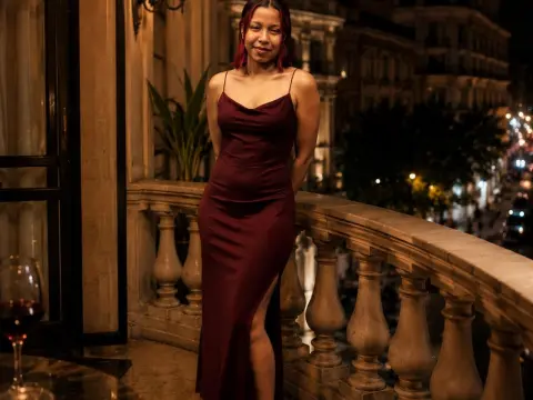 EmilyLovve poses in a long, dark red satin dress with a high slit while standing on a stone balcony overlooking a city street at night.