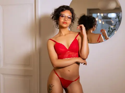 KiraSantisabel poses in red lace lingerie and glasses, showing her dark curly hair and midriff against a white wall and door.