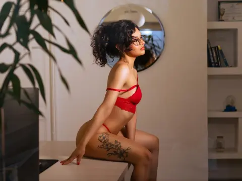 KiraSantisabel poses in red lace lingerie, showing her curly dark hair and hip tattoo while sitting on a white table in a room with a mirror.