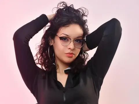KiraSantisabel poses with her hands in her dark curly hair, wearing glasses and a black long-sleeved zip-up top against a pink background.