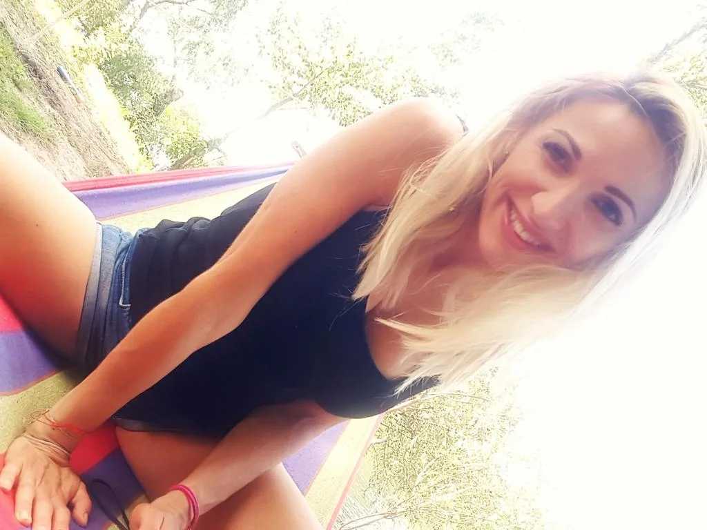 SpecialModelXXXX — Smiling blonde woman in a black tank top and denim shorts relaxing outdoors in a sunny garden setting.