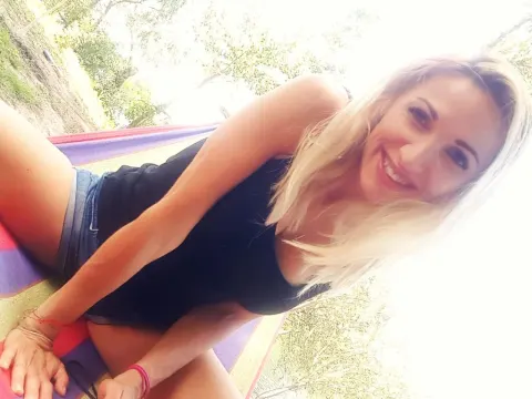 SpecialModelXXXX — Smiling blonde woman in a black tank top and denim shorts relaxing outdoors in a sunny garden setting.