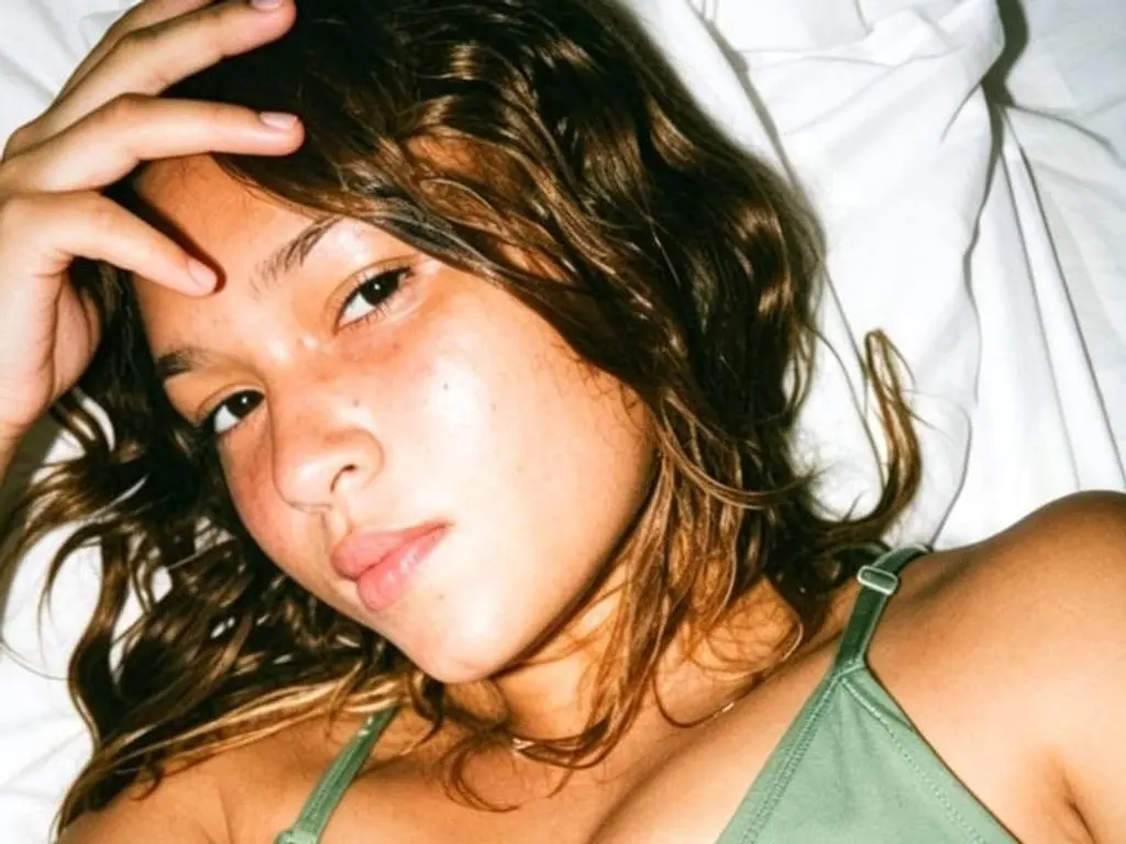 Cataleya_Rouge lying in bed with her hands in her wavy brown hair, wearing a light green bra or bikini top.