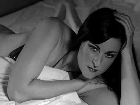MsGennyRock — MsGennyRock lies on a white bed sheet, looking at the camera with her dark hair falling over her face.