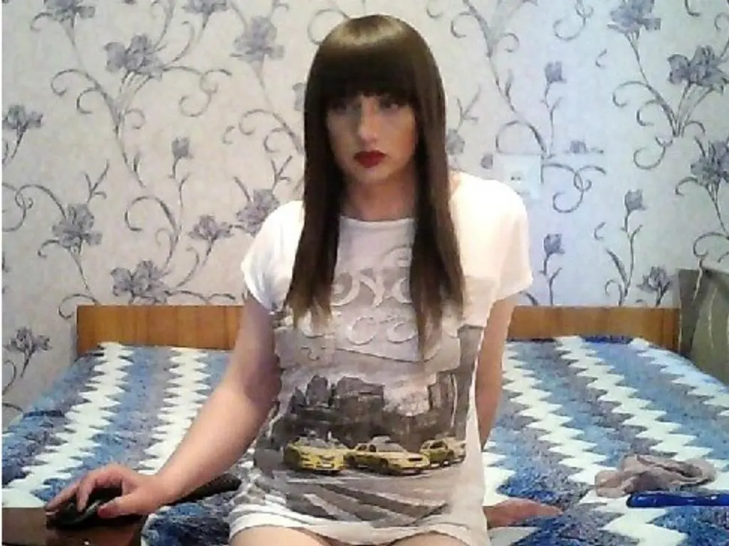 betty3791 has long brown hair with bangs and wears a white t-shirt while sitting on a blue and white patterned bed.