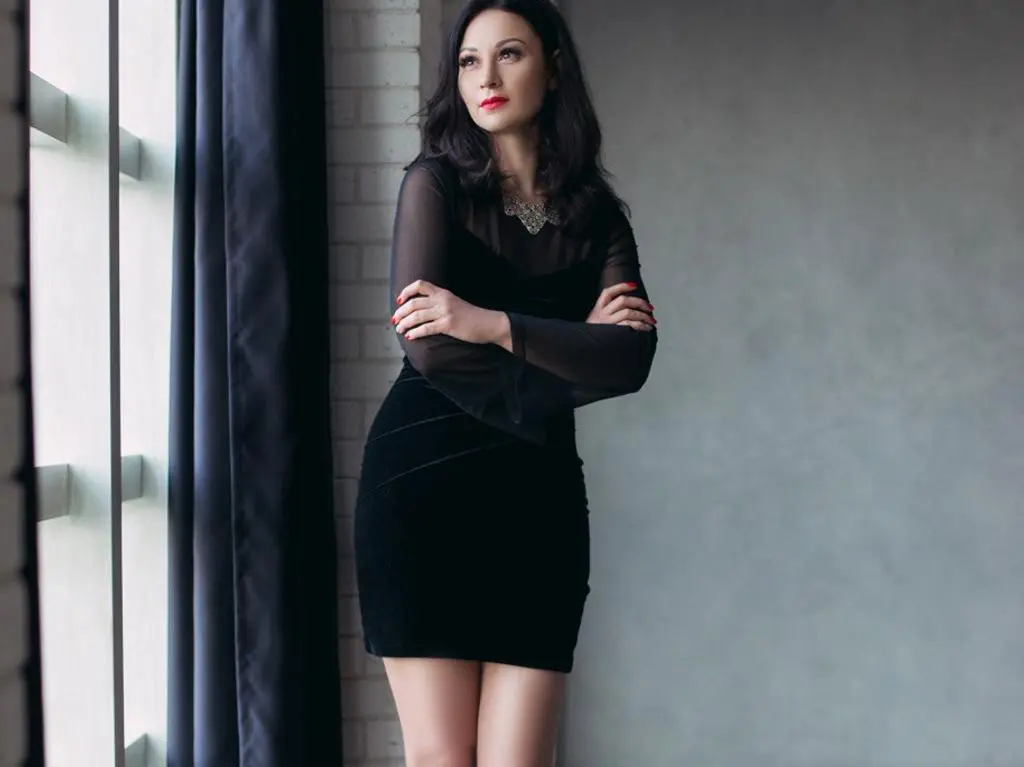 Kethlyne poses in a short black dress with long sheer sleeves and a silver necklace, standing near a window with dark curtains.