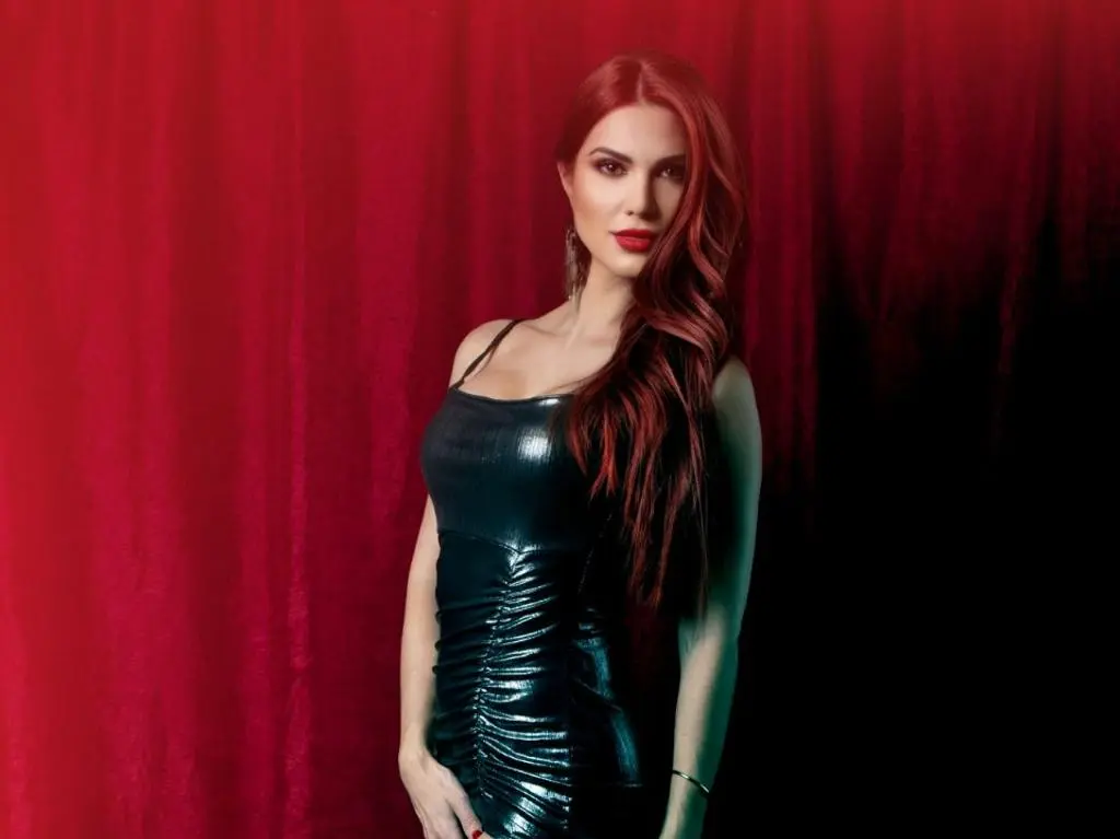AdaGoddess poses in a tight black leather dress against a red velvet curtain, showing off her long red hair and red lipstick.