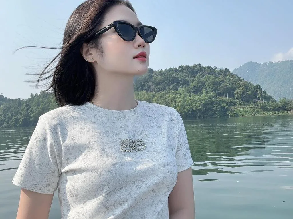JulySex — JulySex wears black sunglasses and a white patterned t-shirt while posing outdoors by a lake and green mountains.