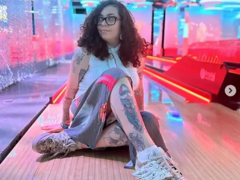 BrazilianHot, wearing glasses and a white top, sits on a stage with her tattooed arms and legs visible, wearing a grey skirt and white sneakers.