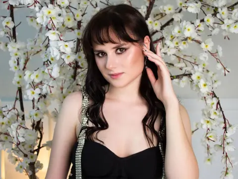 SophiaNoire poses with her hand in her dark hair, wearing a black top against a background of white flowers.