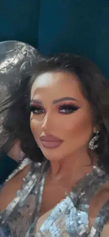 Assyya — Assyya poses with long dark hair and heavy makeup, wearing a sparkly silver sequined top.