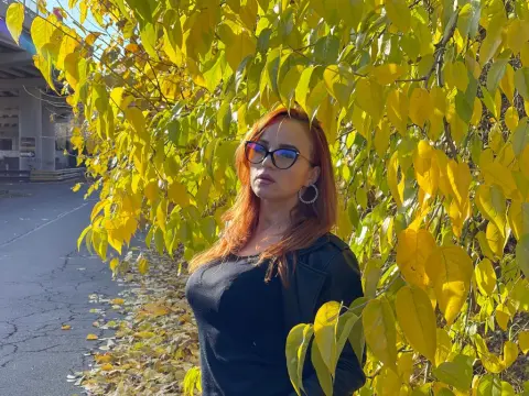 ToxiC_Game — ToxiC_Game is a redhead wearing glasses and a black top, posing in front of bright yellow autumn leaves.