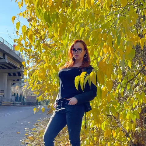 ToxiC_Game — ToxiC_Game stands outdoors in front of bright yellow autumn leaves, wearing glasses, a black long-sleeve top, and dark jeans.