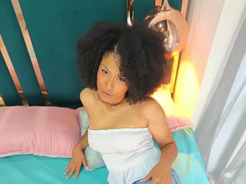Milubrunnete69 sits on a bed wearing a white tube top and a black choker, looking at the camera with her dark curly hair down.