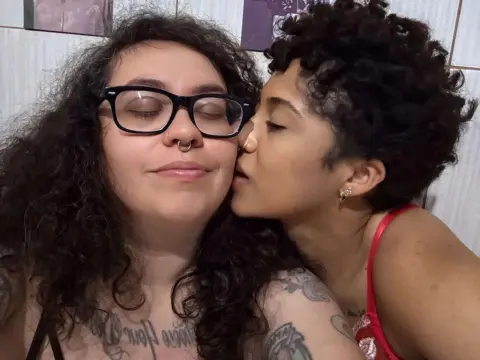 BrazilianHot kisses the cheek of a woman with long curly hair and black glasses, showing her tattoos and short curly hair.