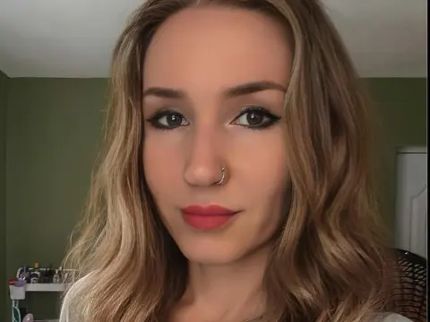 MsPurrfectLovexx looks directly at the camera with blonde wavy hair and winged eyeliner, wearing a light-colored top against a green wall.