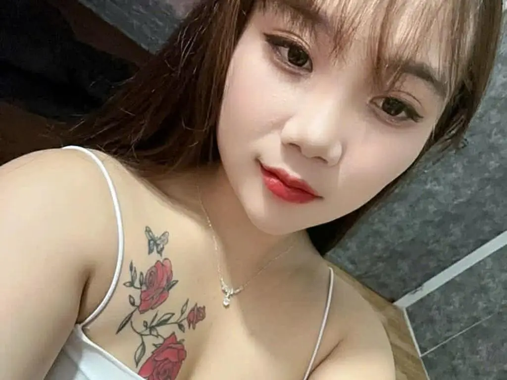Mikabby poses close up, showing her long brown hair, red lipstick, and a large rose and butterfly tattoo on her chest, wearing a white camisole.