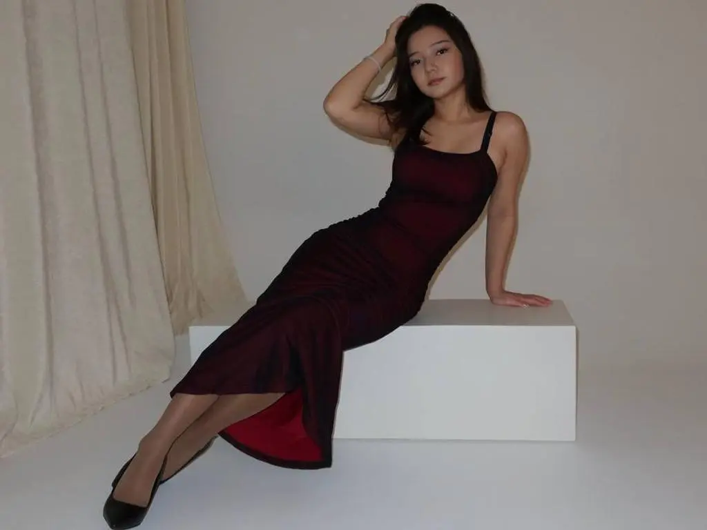 MilanaCares poses sitting on a white block wearing a long, tight dark red dress and black heels against a white background.