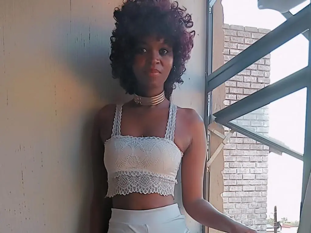 AfricanHotModel poses in a white lace crop top and matching white bottoms, wearing a gold choker and showing her curly dark hair against a brick wall background.