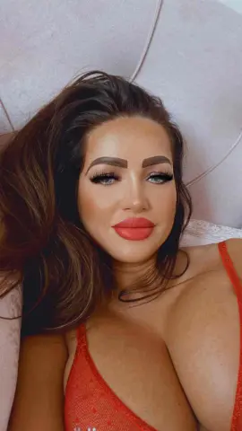 Assyya — Assyya lies in bed wearing a red lace bra, looking at the camera with long hair and heavy makeup.