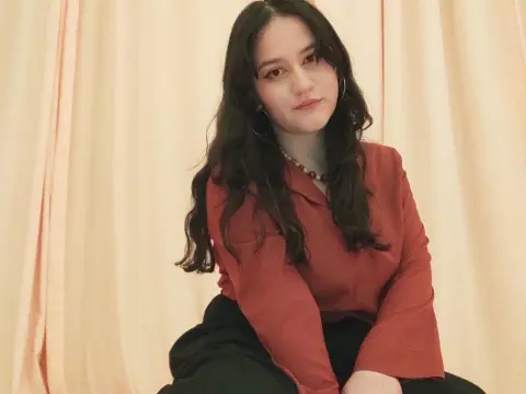 LeylaRosee poses in a red long-sleeve shirt and black pants against a light cream curtain backdrop.