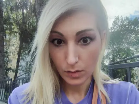 Blonde woman in a purple shirt with dramatic eye makeup posing outdoors near green trees.