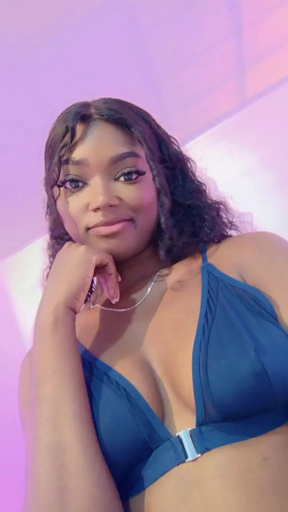 KeisyCollinss — KeisyCollinss poses with her hand under her chin, wearing a blue bikini top and a silver necklace against a soft pink background.