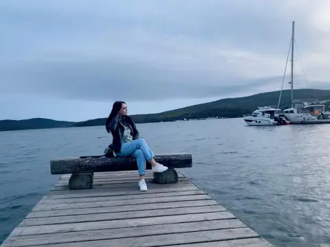 AmelyAngelx — Young woman in casual denim and black jacket relaxing on a wooden pier by a calm lake at dusk.