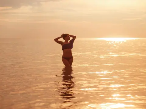 AmelyAngelx — Silhouette of a woman in a bikini standing in the ocean during a golden sunset beach vacation.