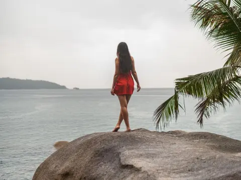 AmelyAngelx — Woman in a red summer dress standing on a large rock overlooking a calm tropical ocean under a cloudy sky.