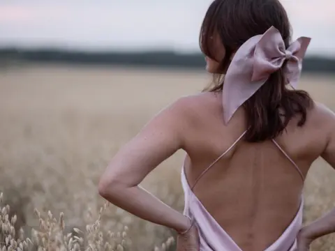 BestHottieEver — Woman in a pink satin backless dress with a large bow hair accessory standing in a dreamy wheat field at dusk.