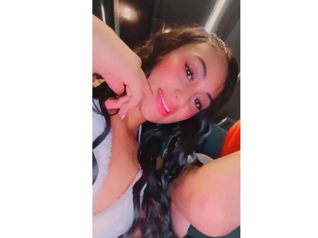 AngelinaStarSS993 smiles while touching her face, showing her long wavy dark hair and white top in a close-up selfie.