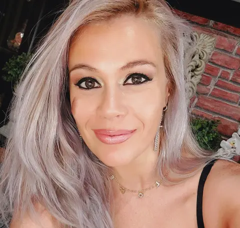 Smiling woman with ash blonde hair and glamorous eye makeup wearing a gold necklace outdoors.
