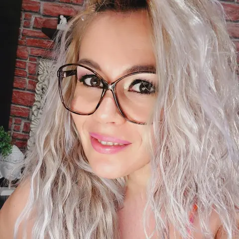 DevilMindXXX — Smiling woman with wavy blonde hair and stylish glasses in a cozy room with a brick wall background.