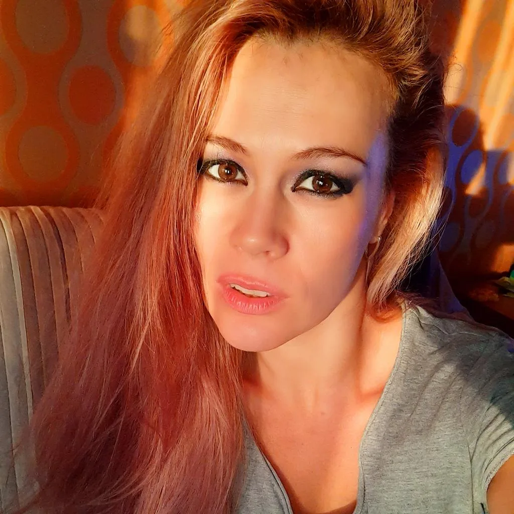 DevilMindXXX — Redheaded woman in a grey t-shirt looking at the camera in warm, moody indoor lighting.