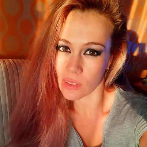DevilMindXXX — Redheaded woman in a grey t-shirt looking at the camera in warm, moody indoor lighting.