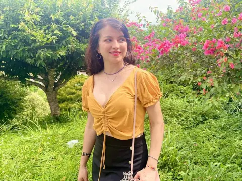 Deb0rahW0nka_ smiles in a yellow top and black skirt while standing outdoors near pink flowers and green trees.