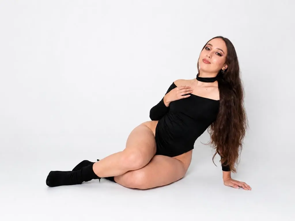 GracieMathews poses sitting on a white floor wearing a black long-sleeve bodysuit and a black choker, showing her long brown hair and bare legs.