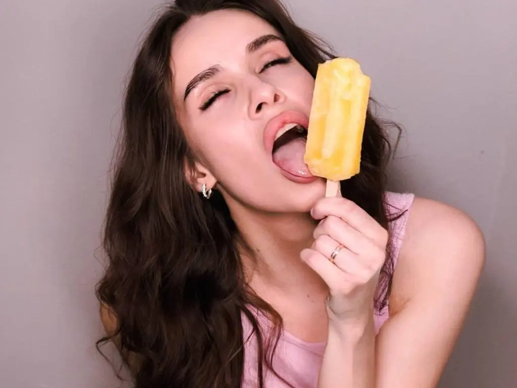 KatyXX licks a yellow popsicle with her tongue, showing her open mouth and long brown hair against a plain grey background.