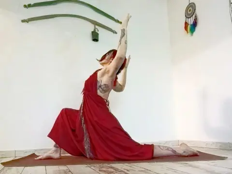 Tattooed woman in a red flowing dress performing a deep yoga stretch on a mat in a bright, peaceful studio.