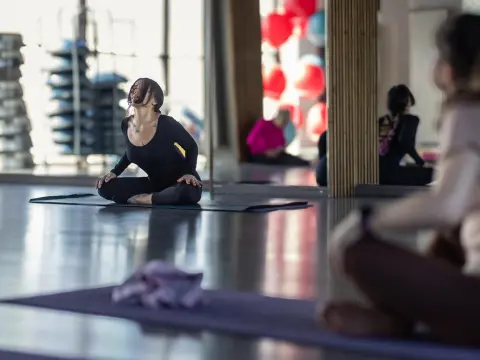 Goddess_Inanna — Goddess_Inanna sits on a yoga mat in a studio, wearing a black long-sleeve bodysuit with her head tilted back and hair falling back.