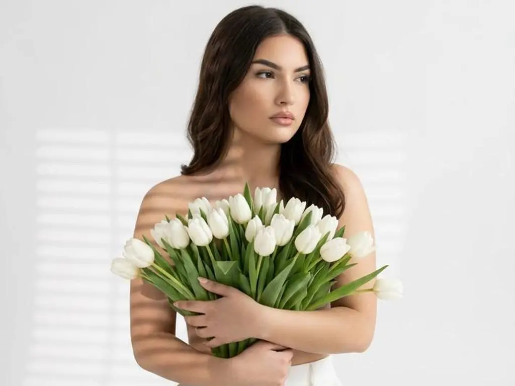 AzzaLeeX holds a large bouquet of white tulips against her bare chest, looking off to the side with long dark hair and a white outfit.