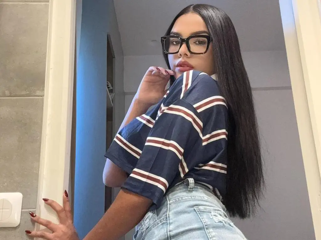 Dailyn4730 poses in a striped blue and white shirt and light denim jeans, wearing black glasses and showing off her long, straight dark hair.