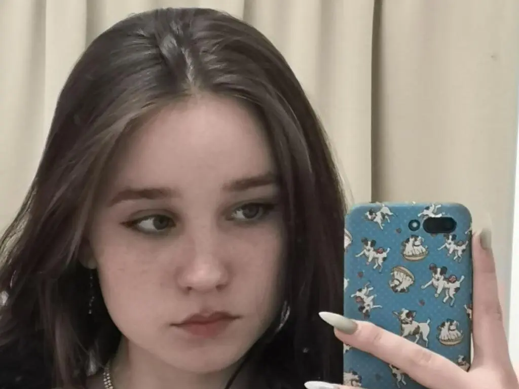 SweetieBlushh takes a close-up mirror selfie, showing her long brown hair and light freckles against a neutral curtain background.