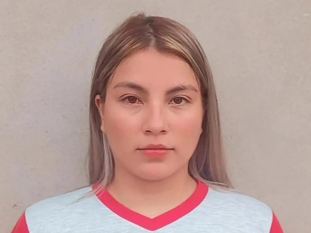 Majo_Sandoval looks directly at the camera with long, light brown hair, wearing a light blue v-neck shirt with red trim against a plain grey wall.
