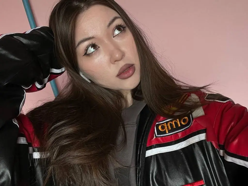 Merida wears a black, red, and white racing jacket and looks up with her long brown hair falling over her shoulders against a pink background.