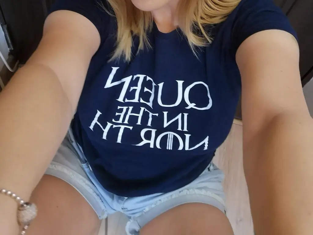 JustKarina — Blonde woman in a navy blue Queen band t-shirt and denim shorts sitting indoors.