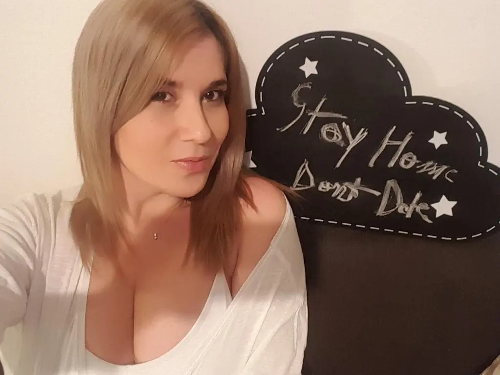 JustKarina — Blonde woman in a white v-neck top sitting in front of a black Stay Home sign in a cozy indoor setting.